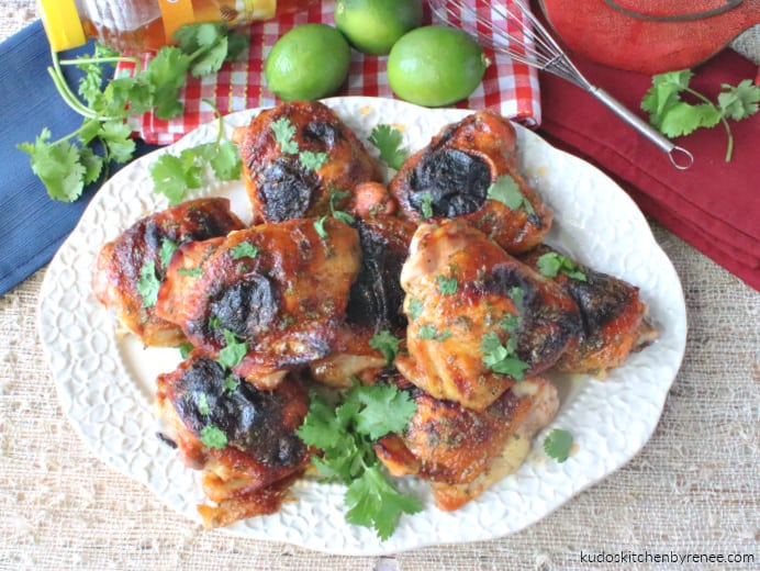 An overhead horizontal photo of a platter of honey lime sriracha chicken with chopped cilantro, a whisk, limes, and napkins in the photo.