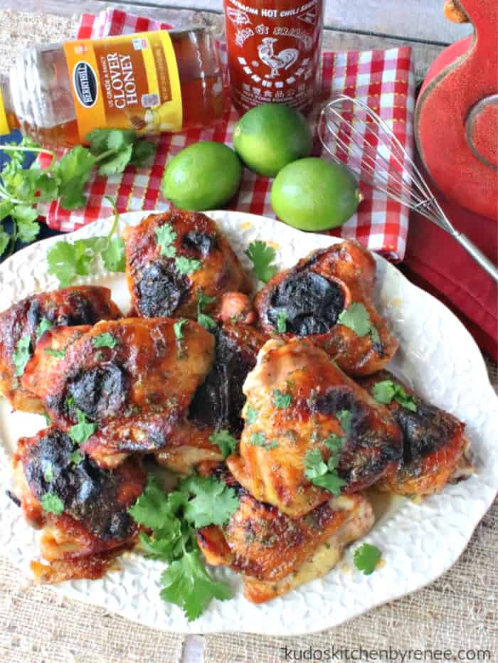 An overhead vertical photo of honey lime sriracha chicken on a platter with a jar or honey, sriracha, limes, and a whisk in the background.