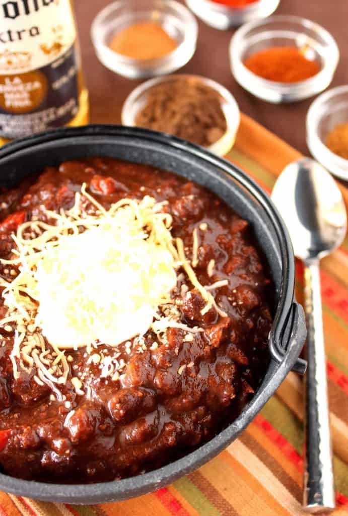 A vertical closeup image of a bowl or turkey mole chili with a spoon on the side and a bottle of beer in the background.