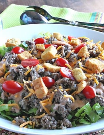 A huge big mac salad in a white bowl with tongs in the background.