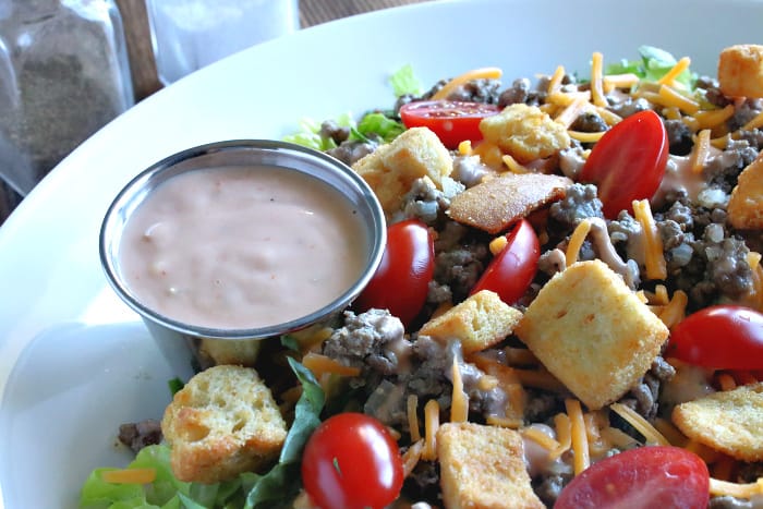 A very close horizontal photo of special sauce on the side of a big bowlful of big mac cheeseburger salad with croutons, cheese, and ground beef.