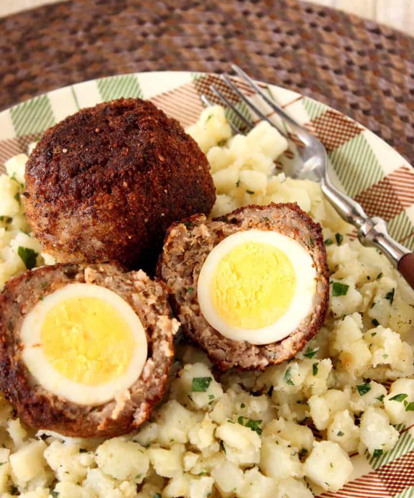 A vertical overhead closeup image of two Scotch eggs on a plaid plate along with some hash brown potatoes and a fork.