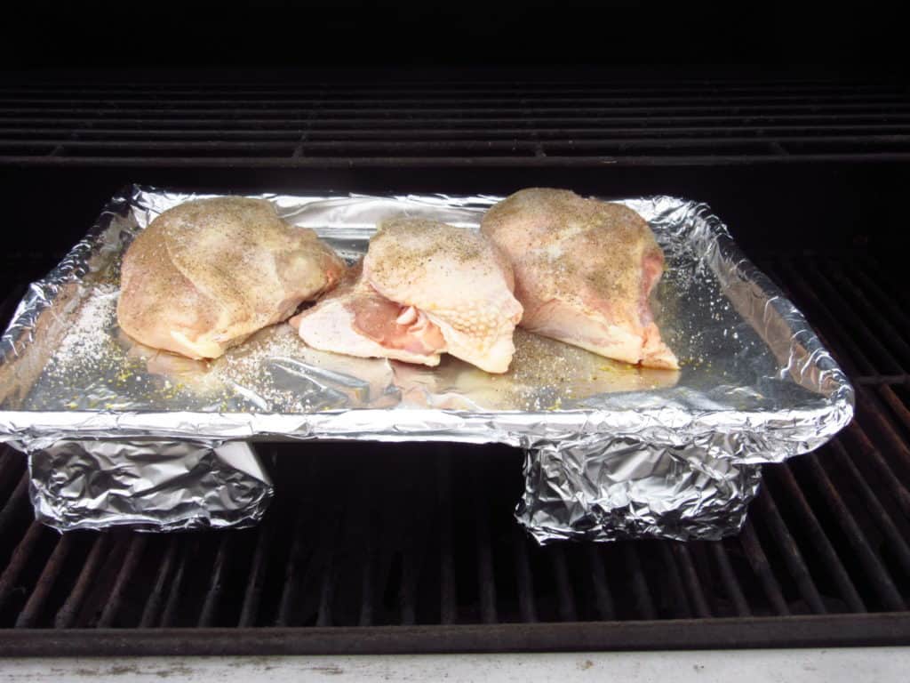 Pieces of uncooked chicken on a baking sheet on an outdoor grill with foil covered bricks.