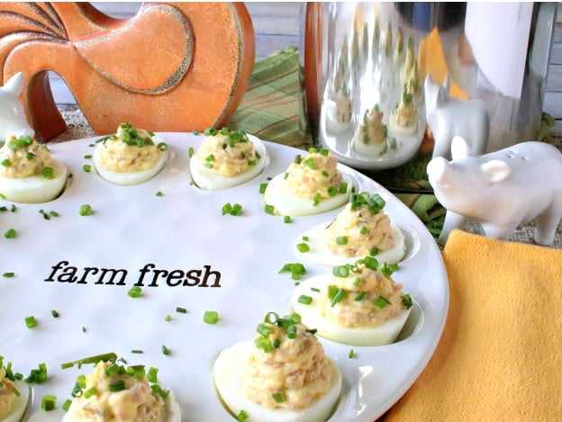 An offset horizontal photo of deviled Dijon eggs with ham and chives on a cute white plate with a sauce pot and a pig salt shaker in the background.