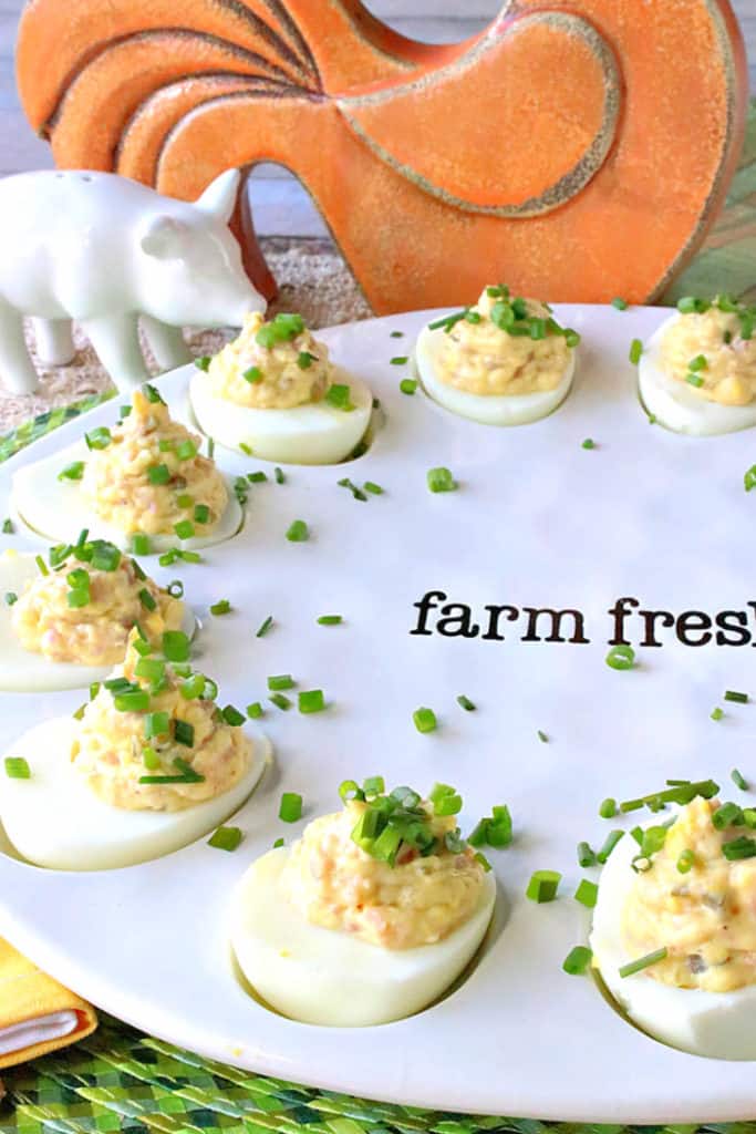 A closeup vertical photo of a tray of deviled Dijon eggs with ham, chopped chives and a ceramic pig salt shaker in the background.