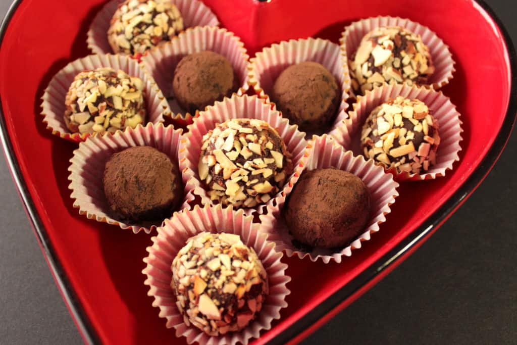 A vibrant horizontal photo of chocolate truffles covered in nuts and coco powder in a red heart shaped dish with paper cups.