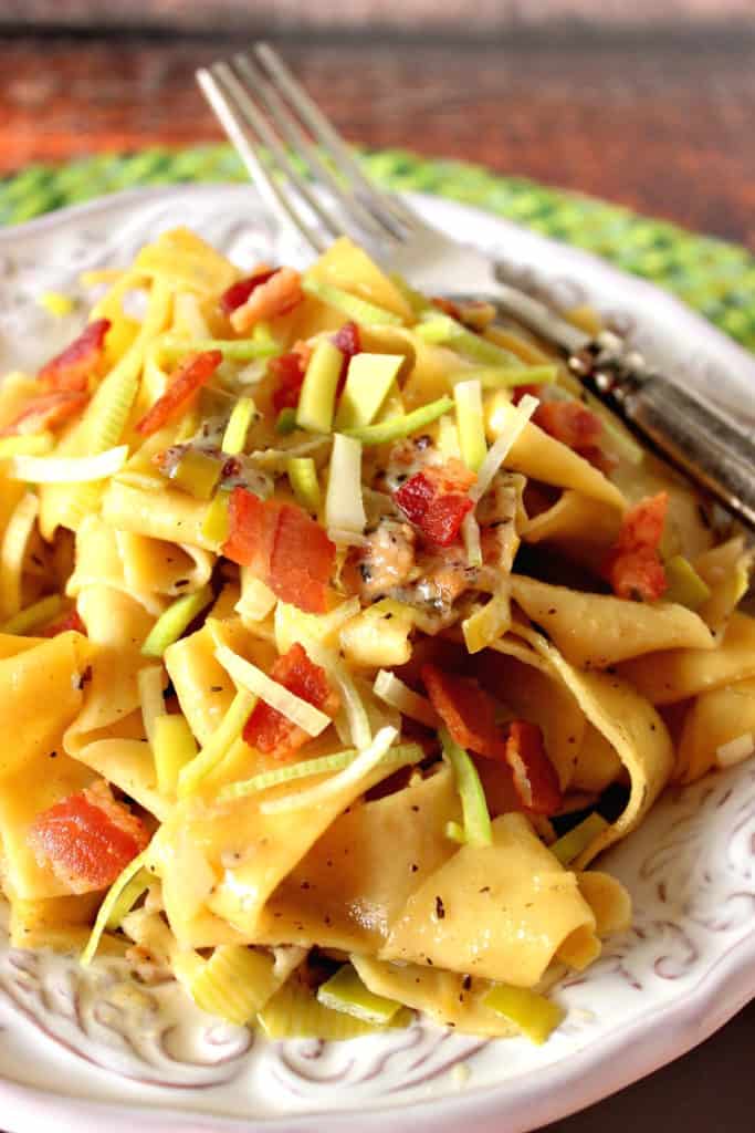 A vertical closeup image of papparadelle pasta on a white plate with leeks, bacon and a fork.