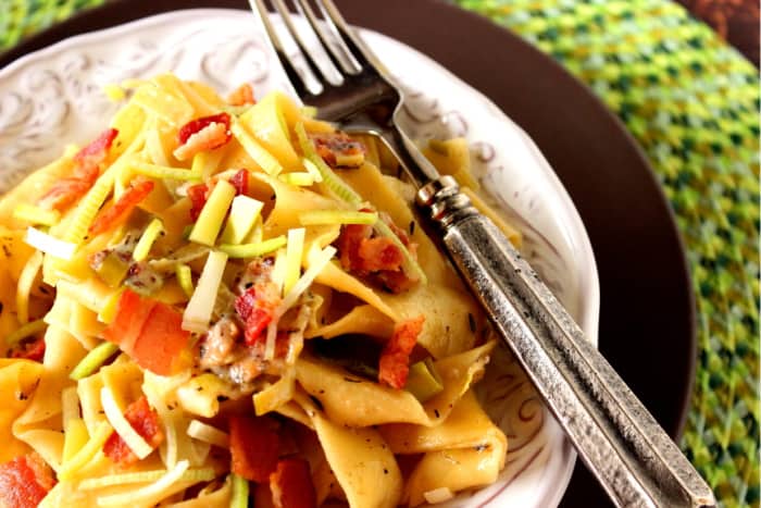 An offset overhead photo of a plate filled with paparadelle pasta with leeks and bacon with a fork and a green placemat.