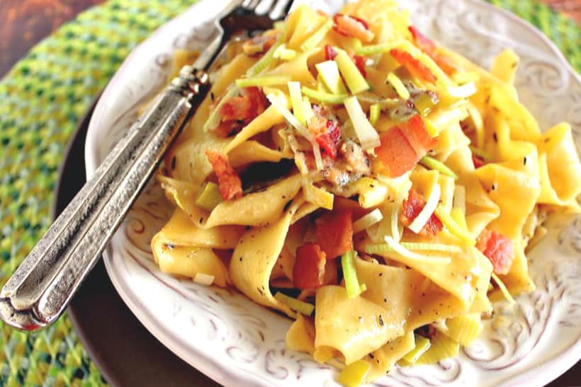 A horizontal photo of a white bowl filled with papparadelle pasta with leeks and bacon on a green place mat with a fork.