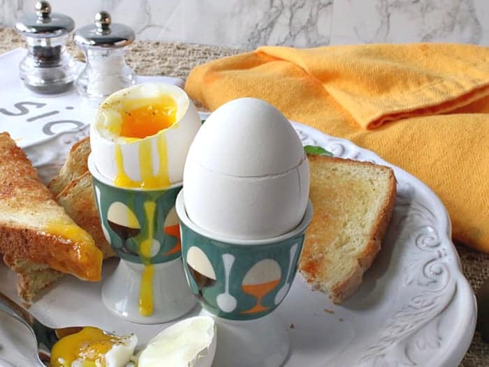 A horizontal photo of soft boiled eggs with runny, drippy yolks in egg cups and salt and pepper shakers in the background.