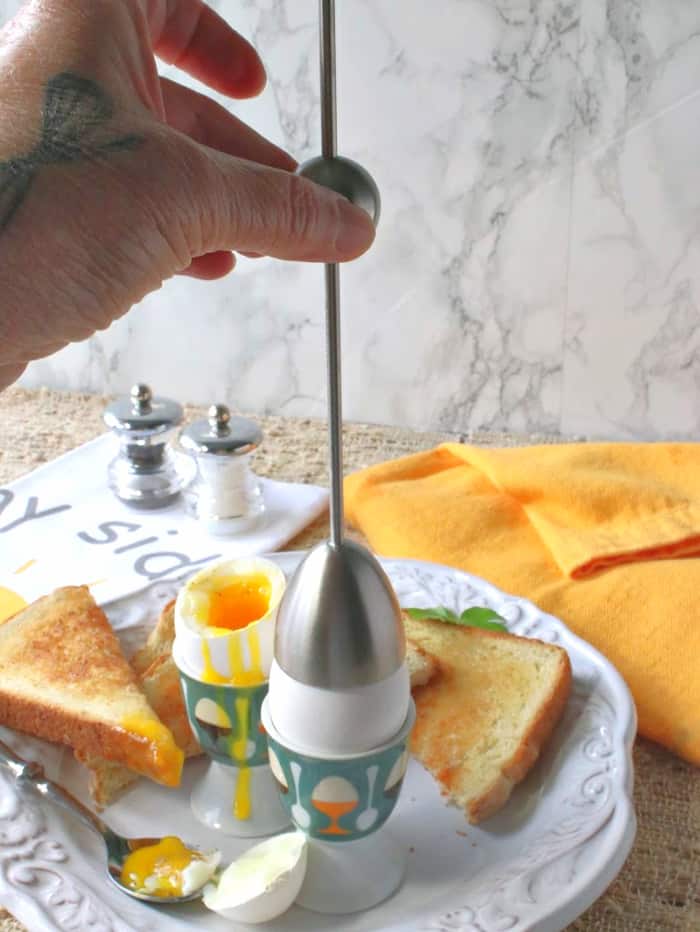 A vertical photo of a hand holding an egg cracker topper on an air fryer soft boiled egg.