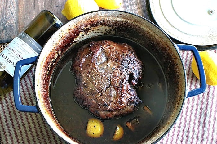 Overhead photo of a deeply browned braised leg of lamb in a Dutch oven with lemon rinds and wine.