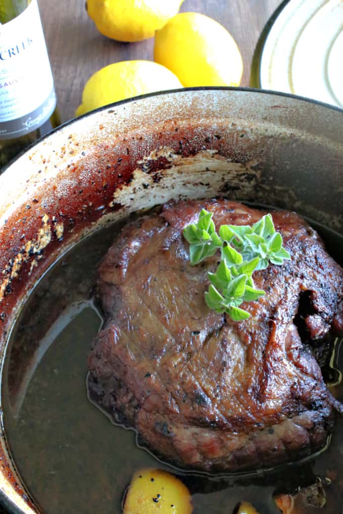 An overhead vertical photo of a beautiful braised leg of lamb in a Dutch oven with oregano sprigs and lemon.