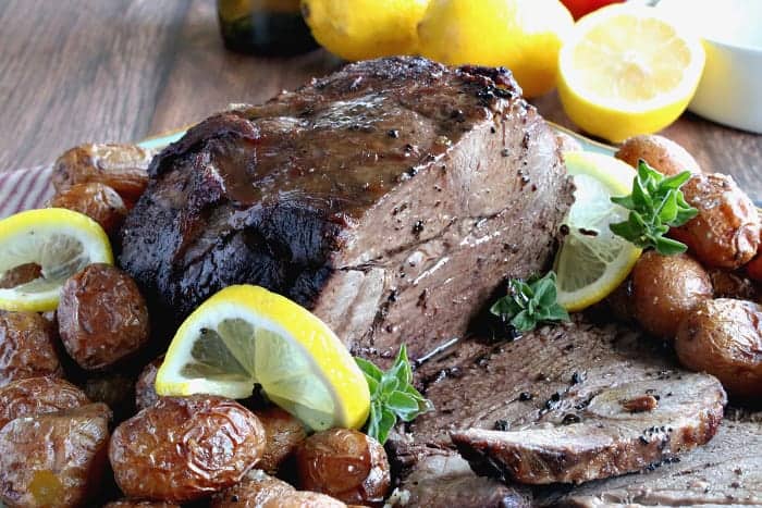 A horizontal photo of a sliced braised leg of lamb with lemons and fresh oregano on a platter with potatoes.