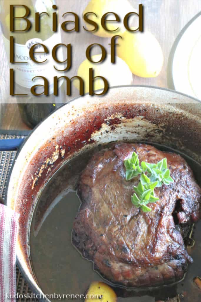 A vertical closeup image of a braised leg of lamb in a Dutch oven with lemon rinds and fresh oregano garnish.