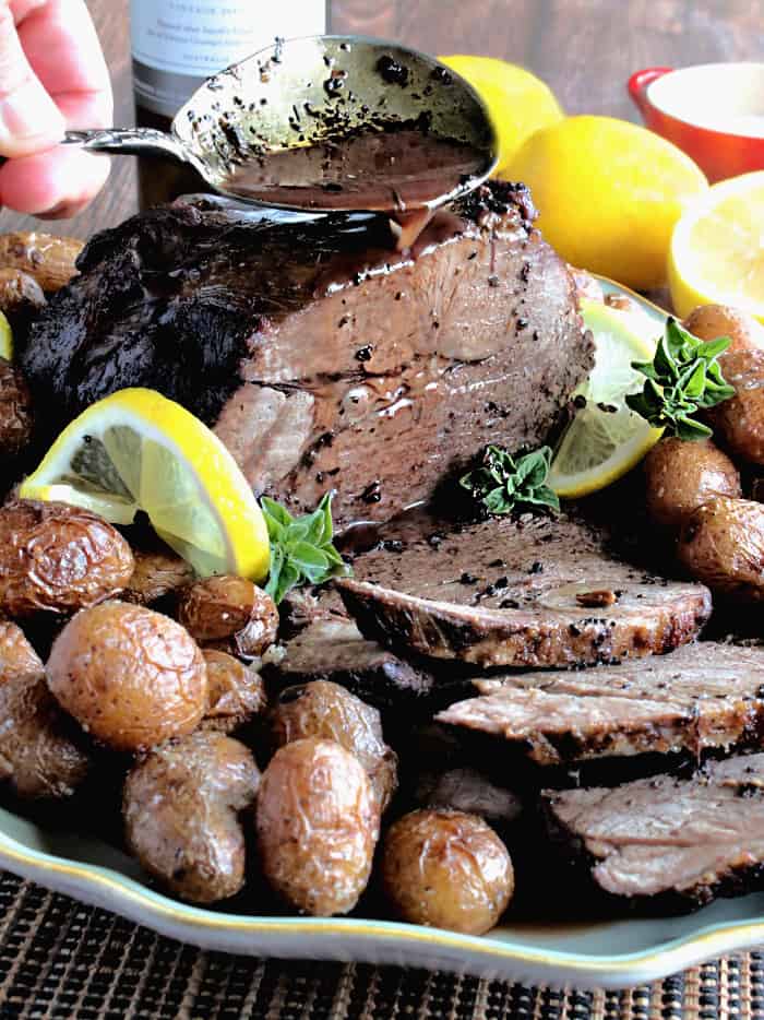 A vertical closeup image of a sliced leg of lamb on a platter with potatoes and lemon slices with a spoon pouring gravy over the meat.