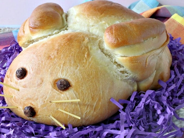 A Easter bunny bread on a white platter with purple Easter grass and colorful napkins.