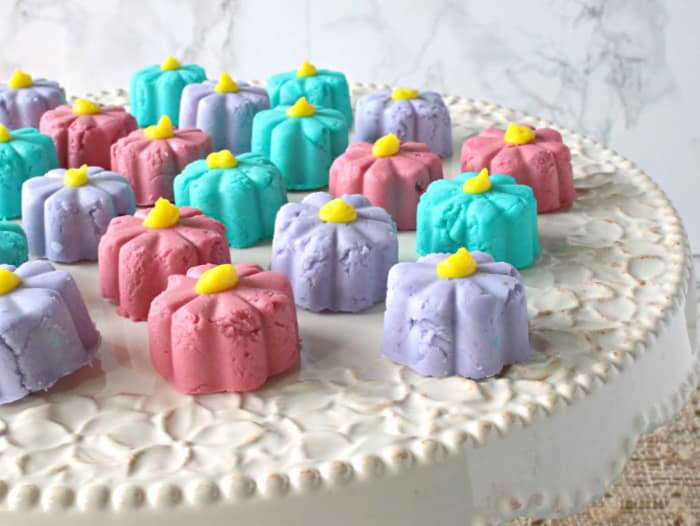 Closeup horizontal photo of pink, aqua, and purple butter mint flowers with yellow centers on a decorative white cake plate.