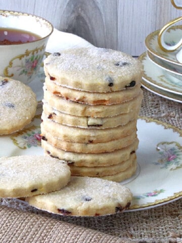 A stack of Orange Cardamom Biscuits on a pretty plate with a cup of tea in the background.