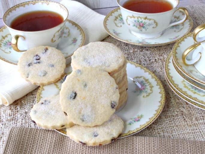 A stack of orange cardamom cookies on a china plate with tea cups in the background.