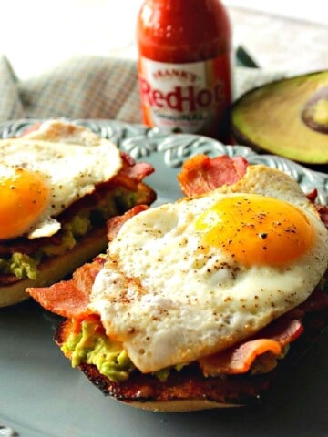 Avocado Toast with Bacon and Egg on a blue plate with hot sauce in the background.