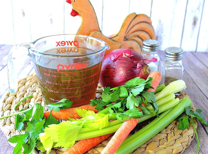 A glass measuring cup filled with budget-friendly and healthy homemade chicken stock with carrots, celery, parsley on a woven rattan place mat. 