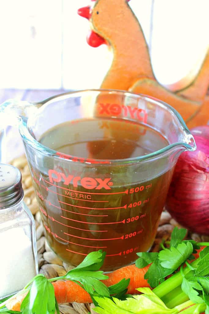 A closeup photo of homemade chicken stock in a glass measuring cup with an onion and a salt shaker on the each side.