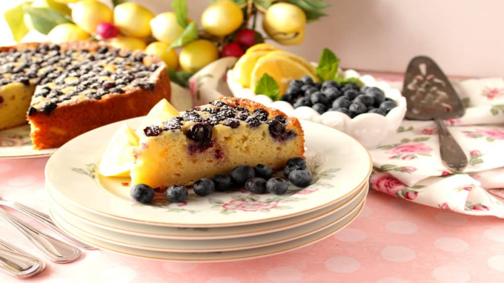 A slice of lemon ricotta cake on pretty plates with a whole cake in the background along with fresh blueberries and lemons