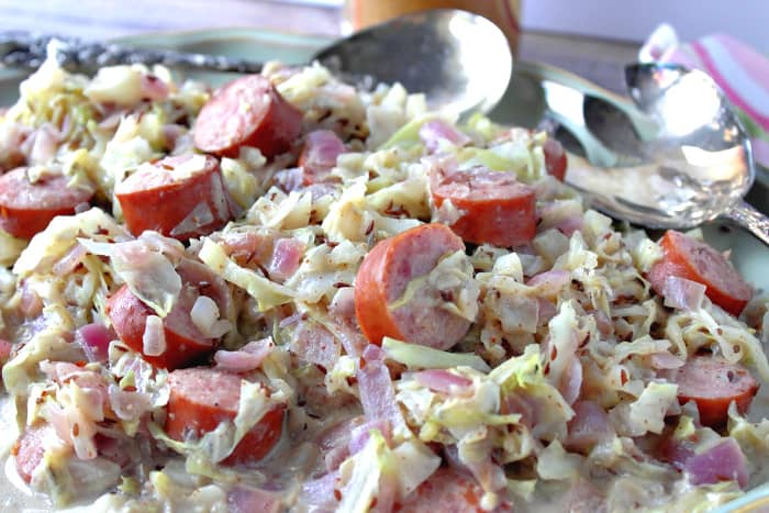Closeup horizontal photo of creamed cabbage with caraway and sausage with silver serving utensils in the background.