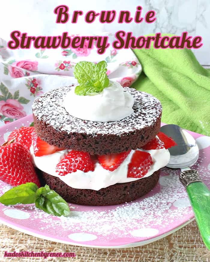 A closeup vertical photo of a brownie strawberry shortcake on a pink polka dot plate with whipped cream, fresh strawberries and mint.