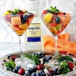 Two martini glasses filled with Boozy Fruit Cocktail with a bottle of gin in the background.