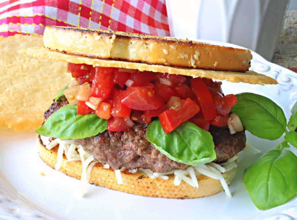 A horizontal photo of a ground beef bruschetta burger on a white plate with red tomato topping and green basil leaves.
