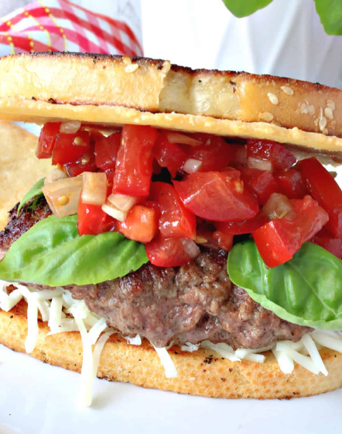 Closeup photo of a ground beef bruschetta burger topped with a Parmesan crisp, tomatoes, and fresh basil leaves.