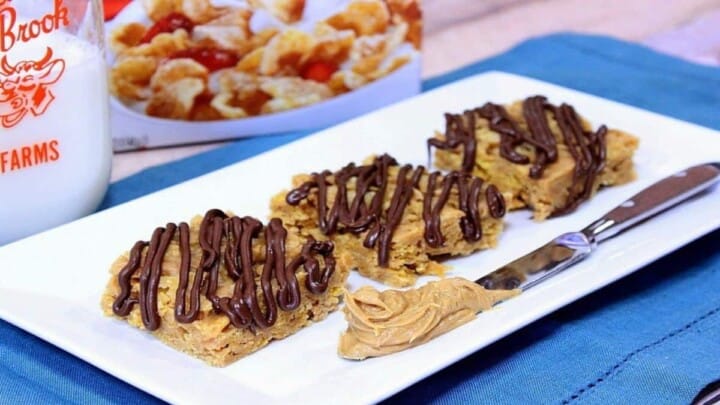 A horizontal photo of a white rectangle plate with tiger paws cereal bars with a chocolate drizzle and a knife with peanut butter on the plate.