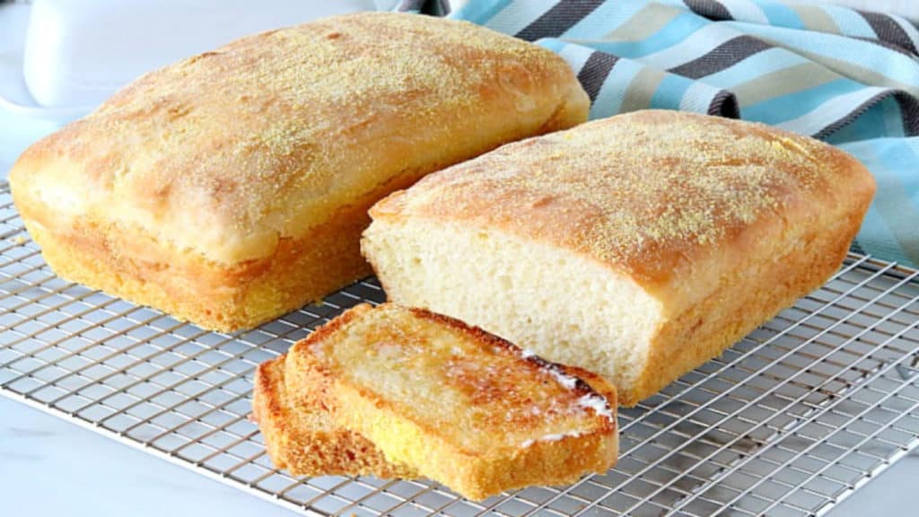 Two loaves of English muffin bread, one sliced and toasted on a wire rack with a blue and brown napkin in the background.