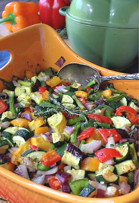 Vertical photo of colorful roasted summer vegetable medley in an orange baking dish with thyme sprigs for garnish, and a serving spoon.
