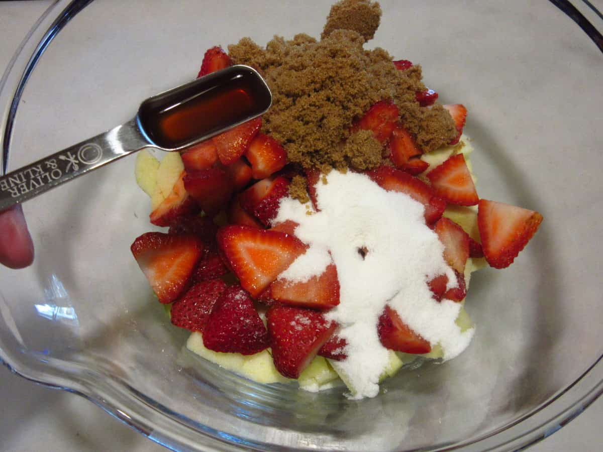 Vanilla extract being added to a bowl with a pie filling of apples and strawberries.