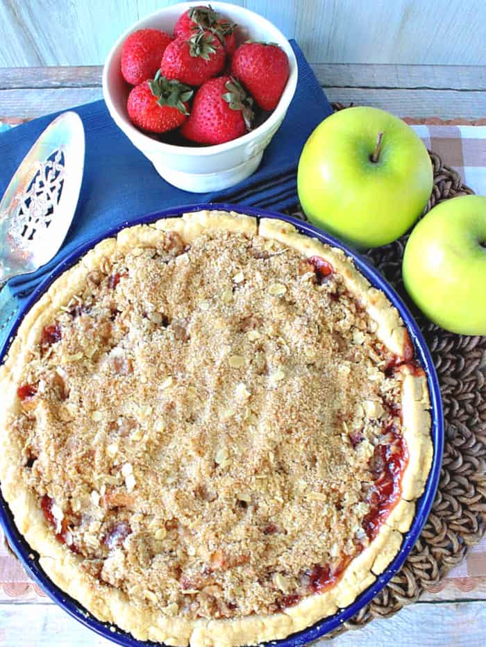 A closeup overhead vertical photo of a strawberry apple pie with streusel topping with a pie server, a dish of strawberries, and two apples.
