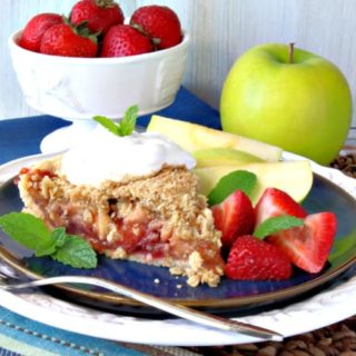 A slice of strawberry apple pie on a blue plate with apple slices and fresh strawberries, mint, and whipped cream.