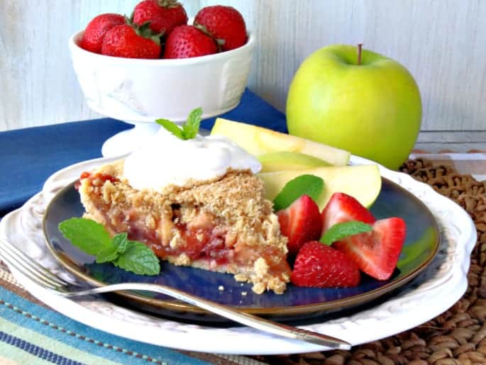 A slice of strawberry apple pie on a blue plate with apple slices and fresh strawberries, mint, and whipped cream.