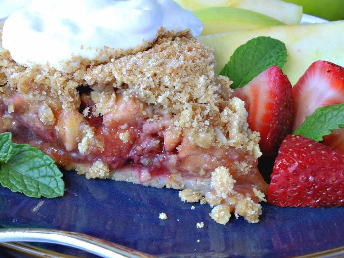 Closeup picture of the inside of a strawberry apple pie with streusel topping, whipped cream, fresh strawberries and mint on a blue plate.