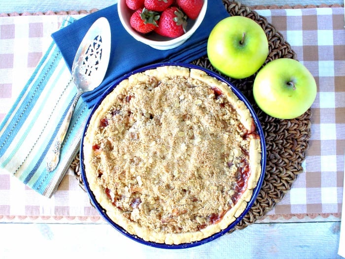 Overhead photo of a strawberry apple pie with streusel topping on a tan and white checked table runner with a silver pie server.