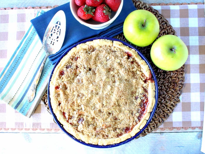 Overhead photo of a strawberry apple pie with streusel topping on a tan and white checked table runner with a silver pie server.