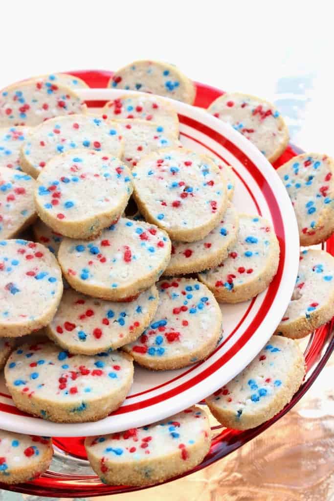 A vertical closeup of a two tiered cookie plate filled with cream cheese cookies with red, white, and blue sprinkles.