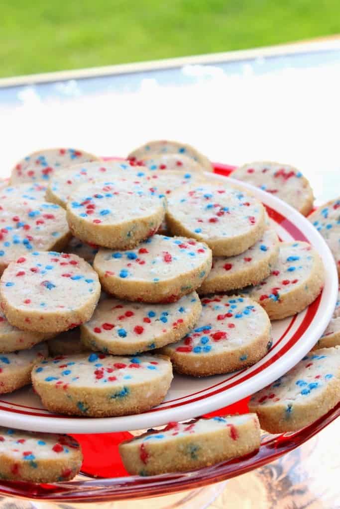 Closeup photo of a plate of cream cheese cookies with red, white, and blue sprinkles with green grass in the background.