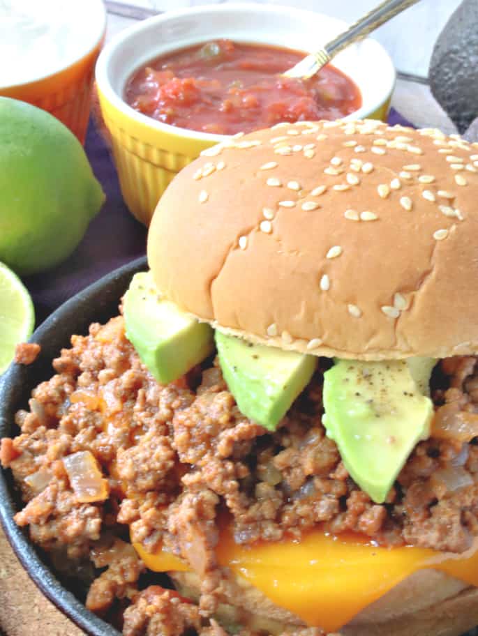 A closeup vertical image of a salsa sloppy joes on a sesame seed bun with limes and salsa in the background.