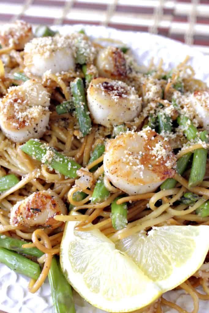 Closeup overhead vertical photo of seared scallops with linguine and asparagus on a platter with lemon slices and breadcrumbs.