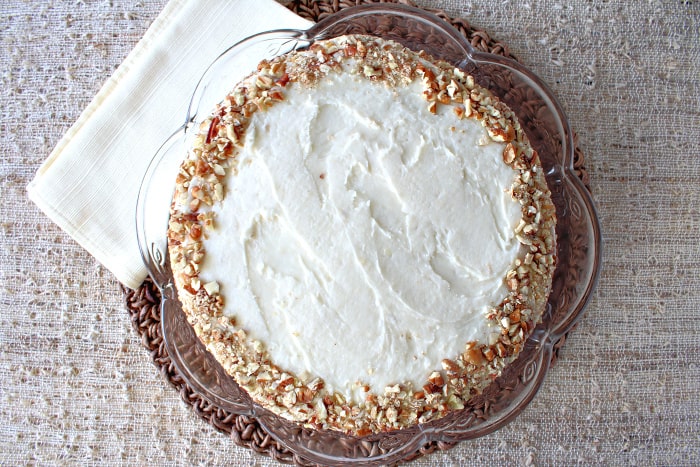 An overhead horizontal photo of an entire banana poppy seed cake on a glass cake plate covered in pecans.