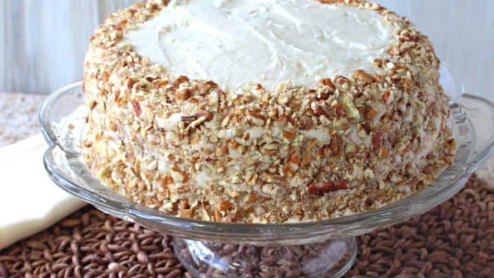 An entire banana poppy seed cake covered in pecans on a glass cake plate.