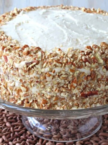 An entire banana poppy seed cake covered in pecans on a glass cake plate.